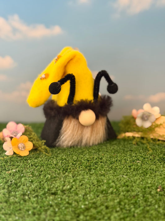 A handmade gnome styled like a bee, featuring a bright yellow hat with black antennae, a fuzzy black and beige body, and a round beige wooden nose, set on artificial green grass with small flowers and a blue sky backdrop. Made by Northern Nomes