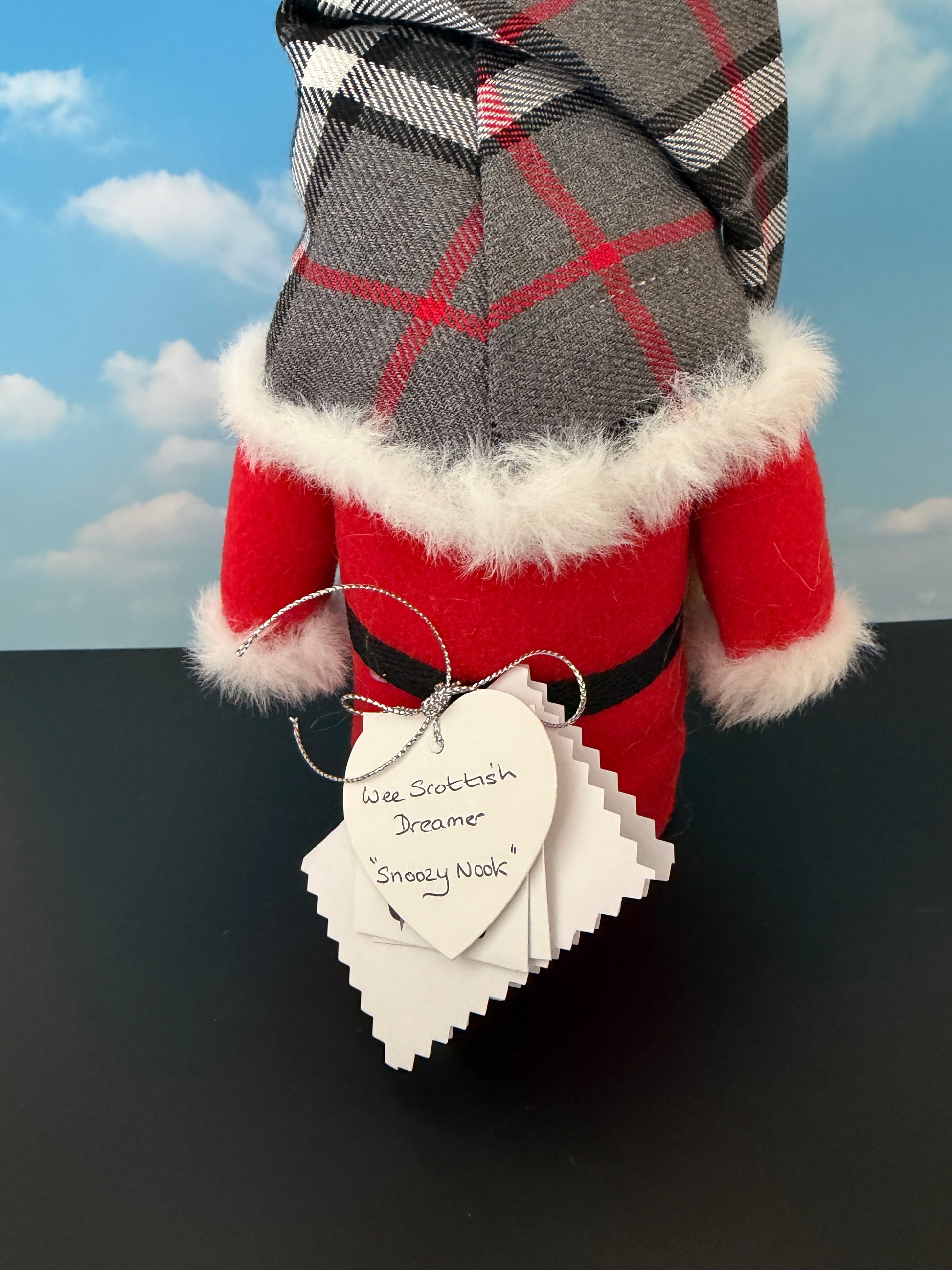 A handmade Scottish Dreamer Gnome shown from the back, wearing a gray and red plaid tartan hat with white faux fur trim, a red body and a white heart-shaped tag that reads 'Snoozy Nook'.