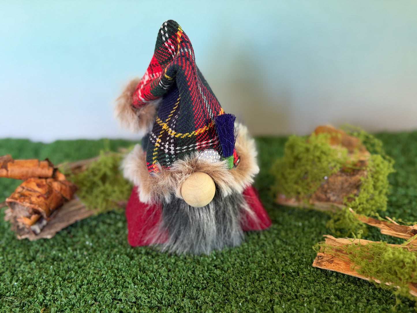 A small decorative gonk figure with a tartan plaid pointed hat, faux fur trim, a gray beard, and a wooden nose, standing on artificial green turf with moss and wood props against a pale blue background. From the Thistlewood Wee Folk Collection by Northern Nomes