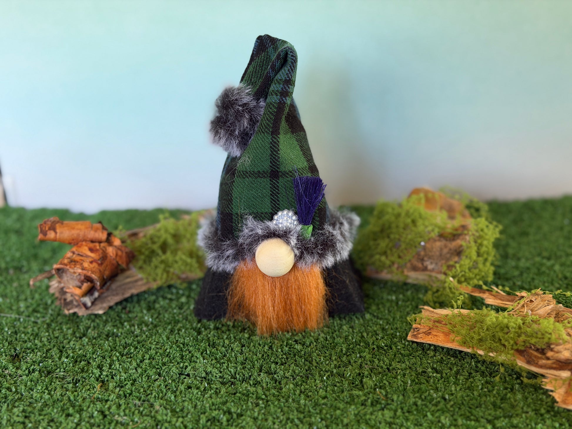 A small decorative gonk figure with a tartan plaid pointed hat, faux fur trim, a ginger beard, and a wooden nose, standing on artificial green turf with moss and wood props against a pale blue background. From the Thistlewood Wee Folk Collection by Northern Nomes