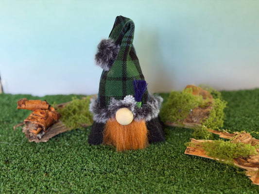 A small decorative gonk figure with a tartan plaid pointed hat, faux fur trim, a ginger beard, and a wooden nose, standing on artificial green turf with moss and wood props against a pale blue background. From the Thistlewood Wee Folk Collection by Northern Nomes
