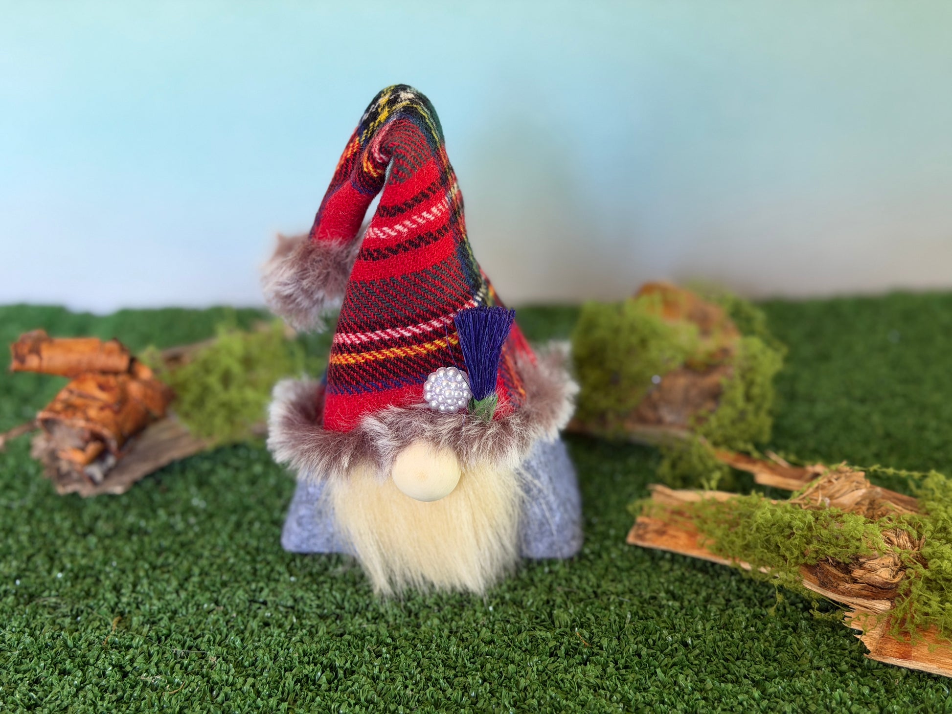 A small decorative gonk figure with a tartan plaid pointed hat, faux fur trim, a blond beard, and a wooden nose, standing on artificial green turf with moss and wood props against a pale blue background. From the Thistlewood Wee Folk Collection by Northern Nomes
