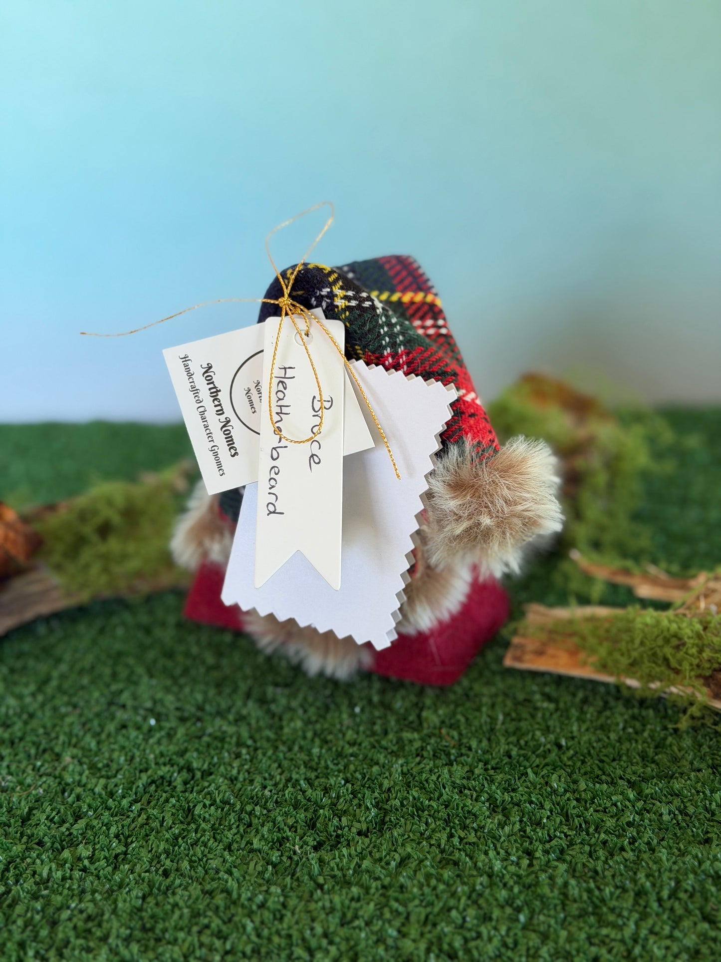 A small handmade gonk figurine dressed in a red and black tartan plaid top hat with a fluffy pom-pom and a red felt body, displayed on artificial green moss with a light blue background. It has hanging tags, including a tag that reads 'Bruce Heatherbeard' and a story description card by Northern Nomes.
