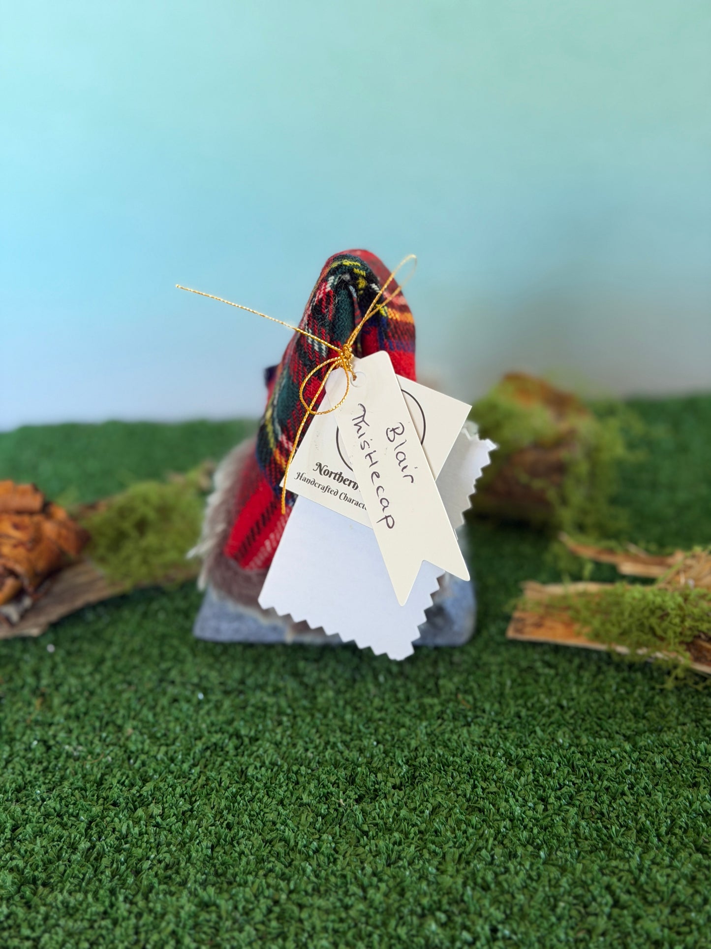 A small handmade gonk figurine dressed in a mixed red tartan plaid top hat with a fluffy pom-pom and a grey felt body, displayed on artificial green moss with a light blue background. It has hanging tags, including a tag that reads 'Blair Thistlecap' and a story description card by Northern Nomes.