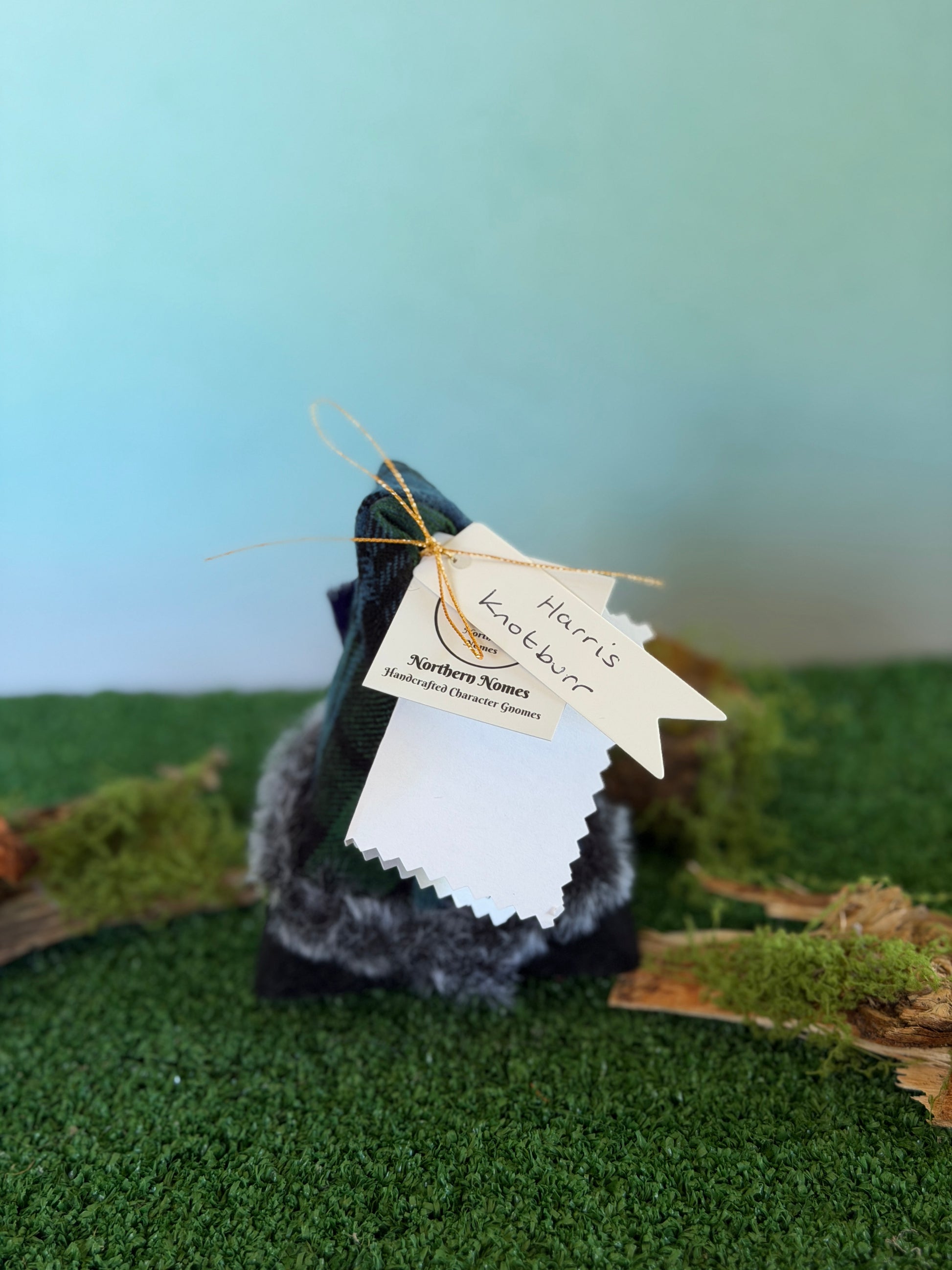 A small handmade gonk figurine dressed in a green and black tartan plaid top hat with a fluffy pom-pom and a felt body, displayed on artificial green moss with a light blue background. It has hanging tags, including a tag that reads 'Harris Knotburr' and a story description card by Northern Nomes.