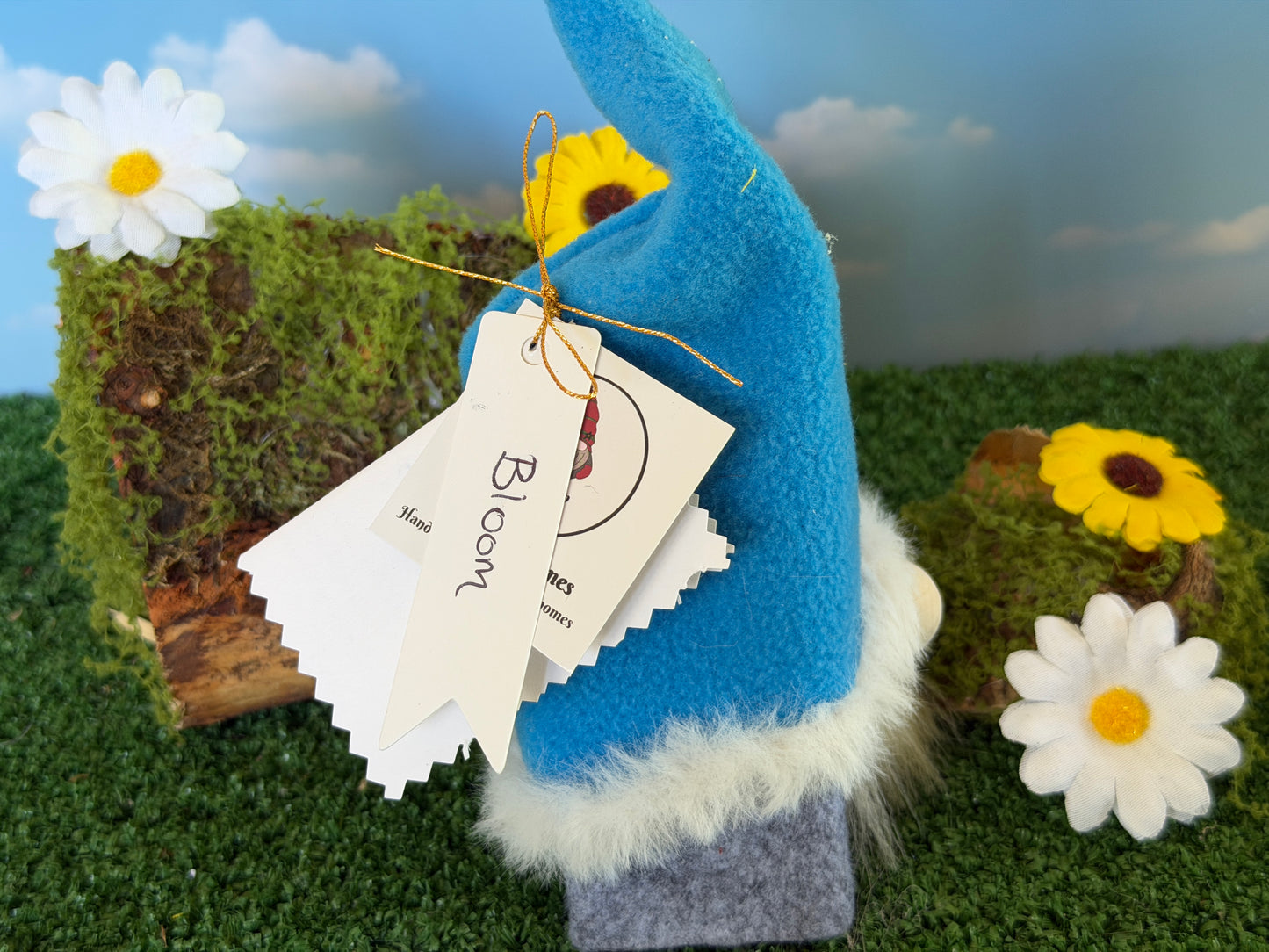 A small blue felt gnome figurine with a white faux-fur trim and a hanging paper tag reading 'Bloom', posed on artificial grass with daisies and a mossy prop against a painted sky backdrop.