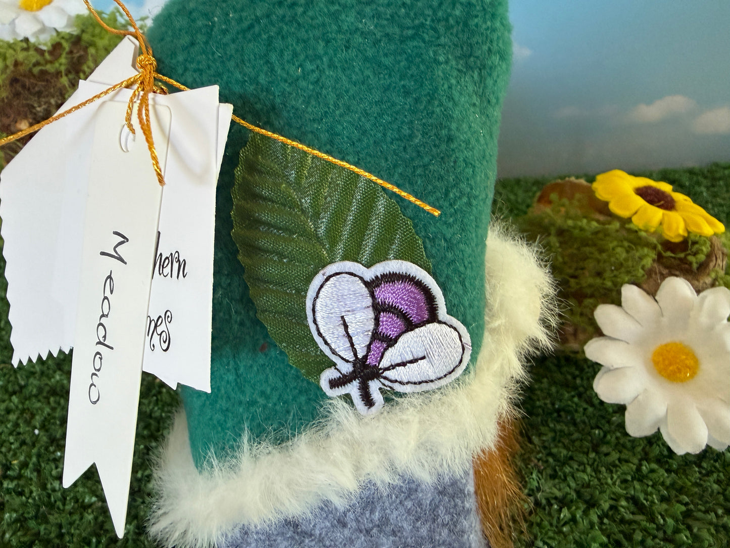 A close-up of a handmade felt gnome hat in teal green with white faux fur trim, featuring an embroidered butterfly patch on the side and a hanging white paper tag.