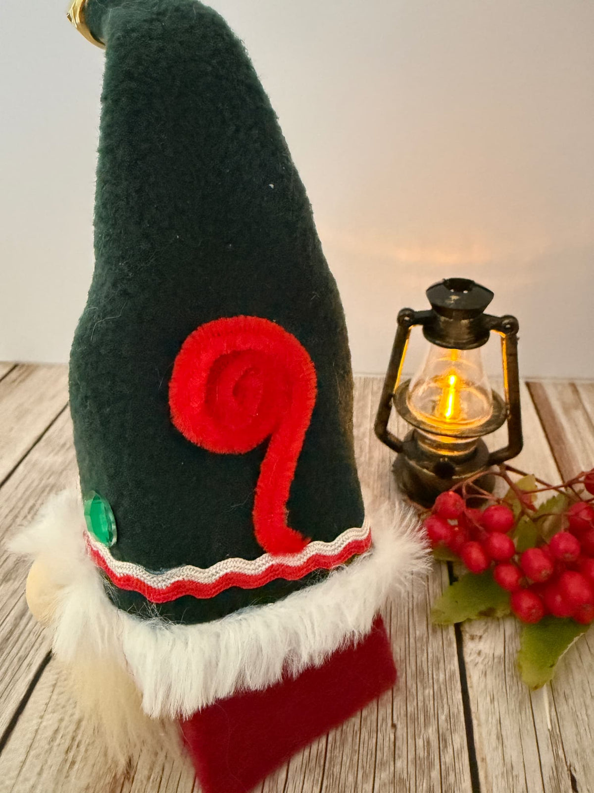 A dark green felt Christmas gnome hat decoration with a red felt swirl, white faux fur trim, and a red brim, displayed on a rustic wooden surface next to a lit lantern and red berries.