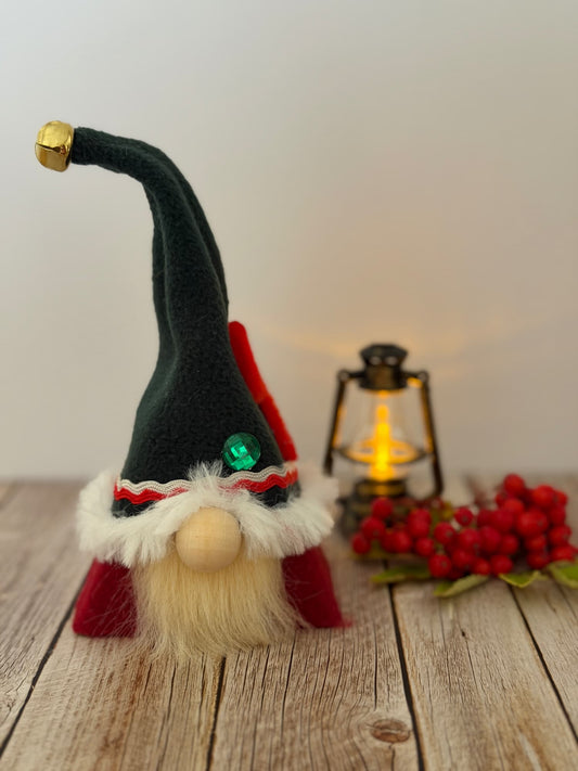A small handmade Christmas elf gnome decoration with a tall dark green hat, white faux fur trim, red clothing, a cream beard, and a green jewel accent, displayed on a wooden surface with a lantern and red berries in the background. From the Festive Folk collection by Northern Nomes.