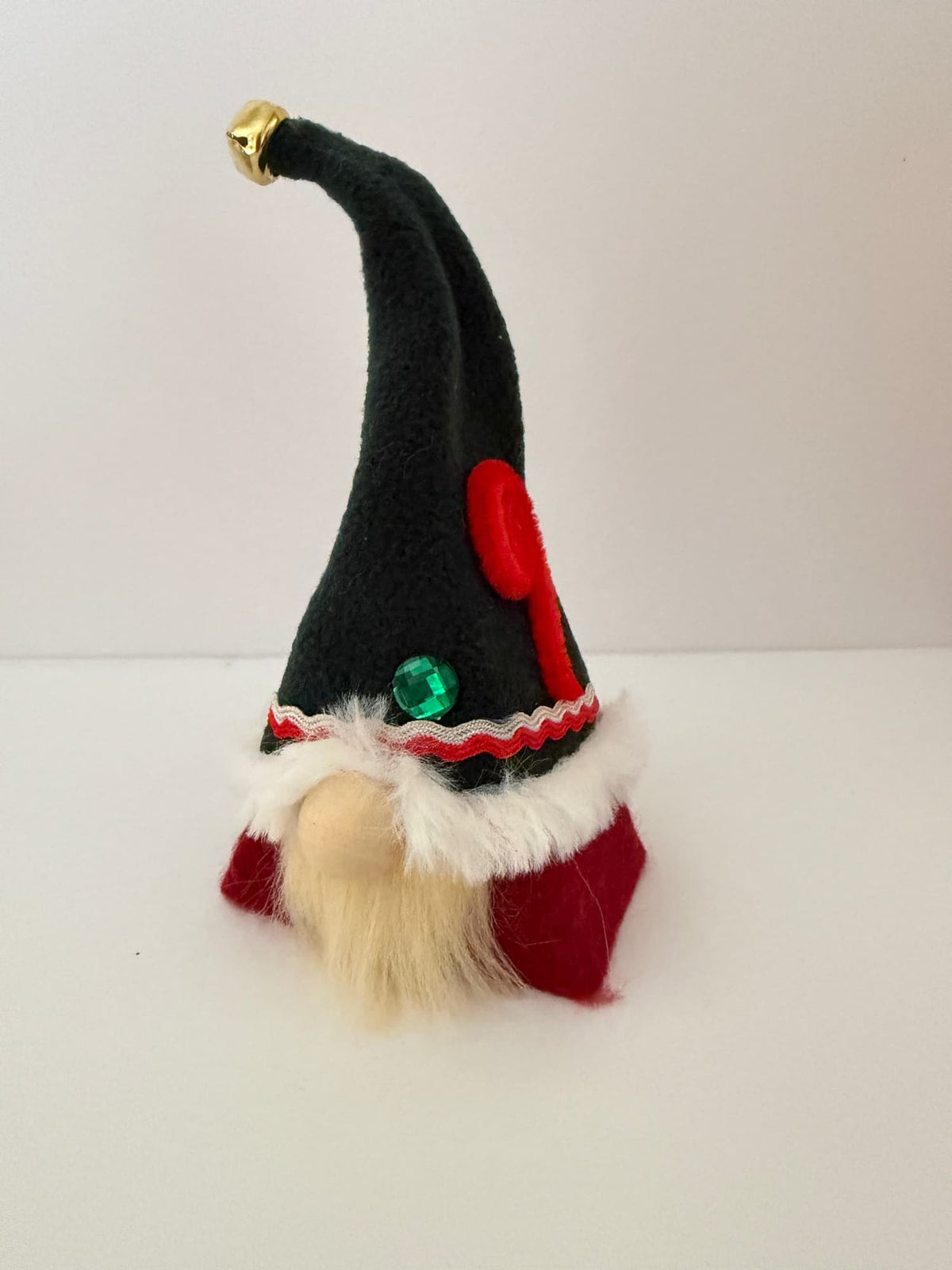 A small Christmas elf gnome ornament with a tall black felt hat tipped in gold, a fluffy cream beard, white faux-fur trim, red clothing, and decorative red and green embellishments, photographed on a plain white background. Handmade by Northern Nomes and part of the festive folk collection