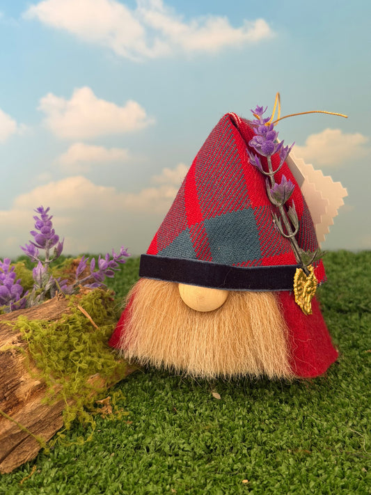 A handmade gnome figurine with a red and blue plaid hat, beige faux fur beard, and a heather sprig with gold accent on his hat, set on artificial grass with a wooden log and a sky backdrop.