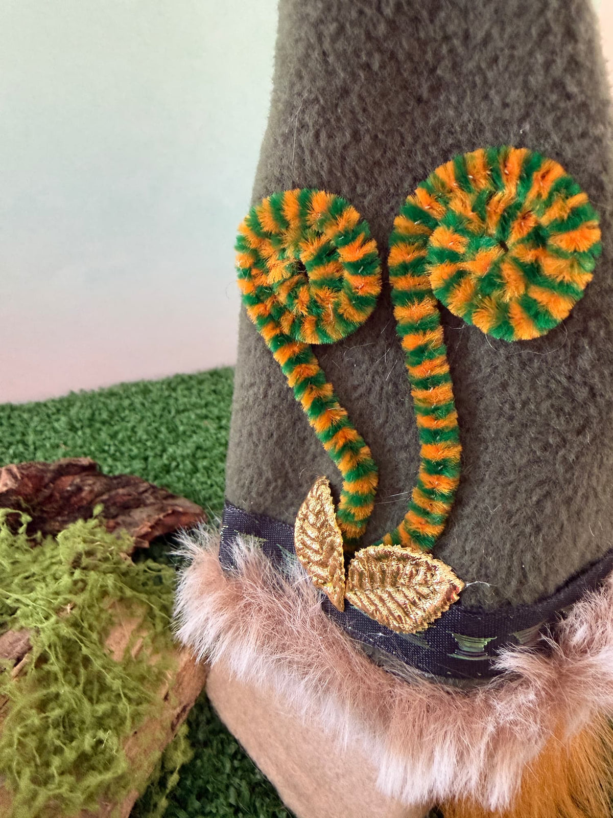 A close-up of a greenish fleece hat with green and orange striped fern style embellishment, a gold leaf applique, and faux fur trim, set against a themed mossy background.
