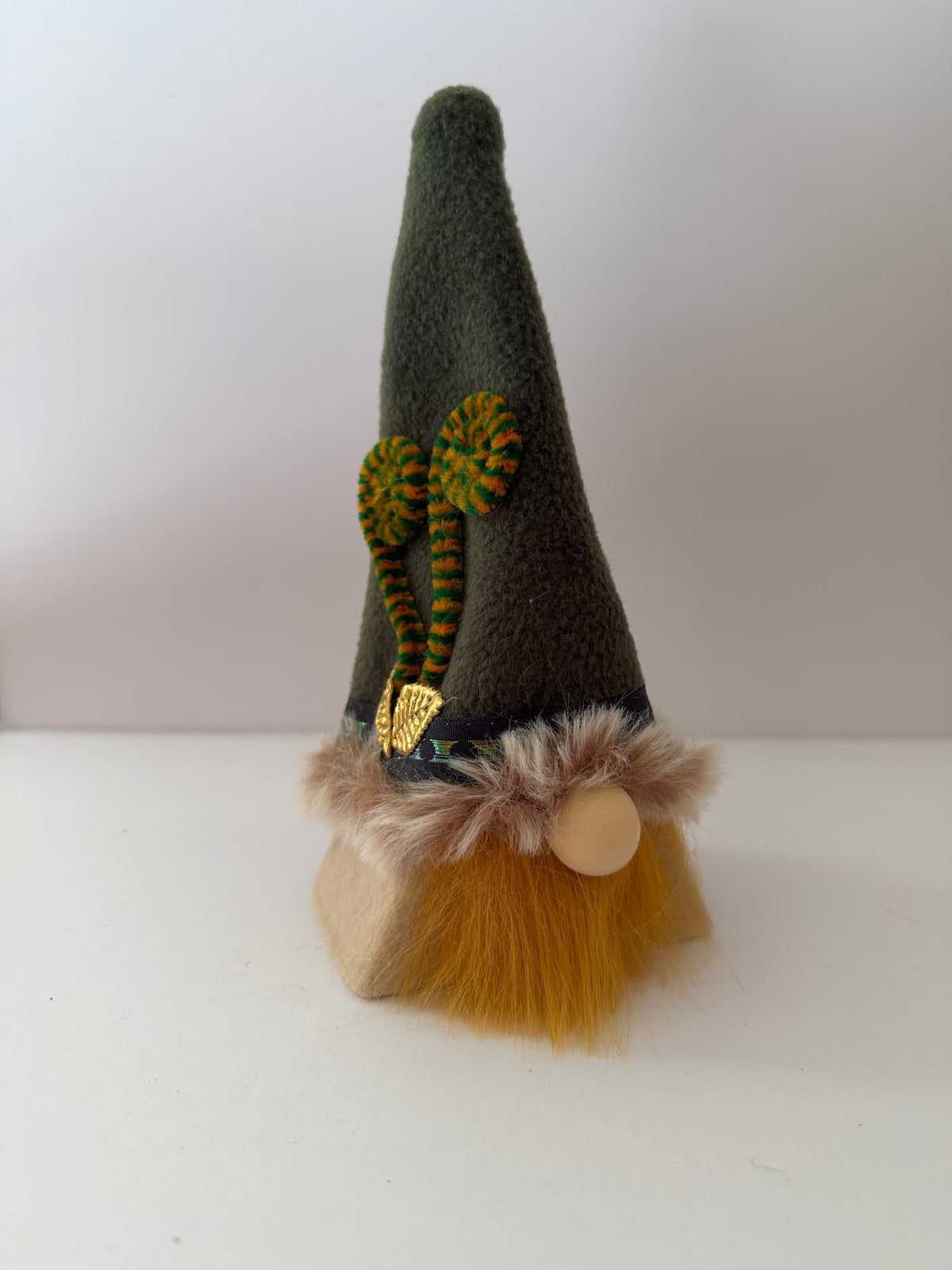 A small gonk with a tall greenish hat, fern styled embellishments, 2 golden leaves, faux fur trim, ginger beard and wooden bead nose, against a white background. Handmade gonk by Northern Nomes. 