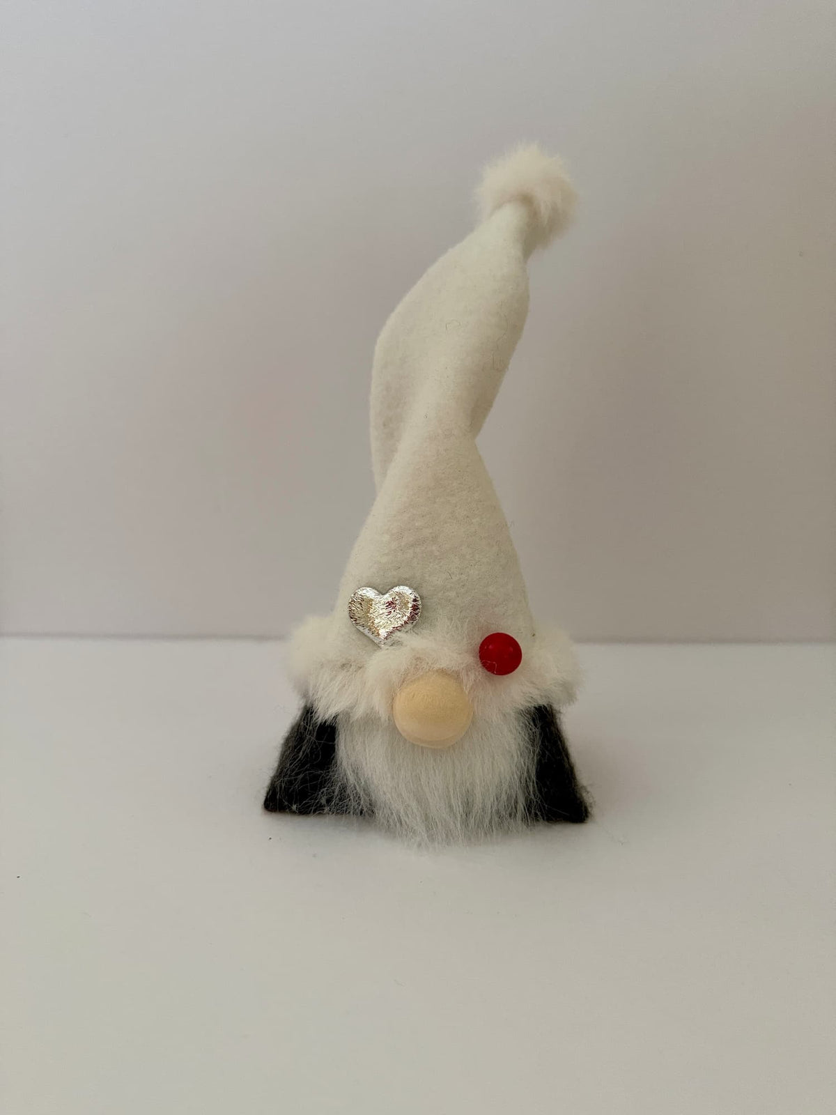A handmade small gnome decoration with a tall white hat, white beard, black felt covered body, a wooden button nose, and a silver heart ornament, standing on a white surface against a light gray background. Made by Northern Nomes.