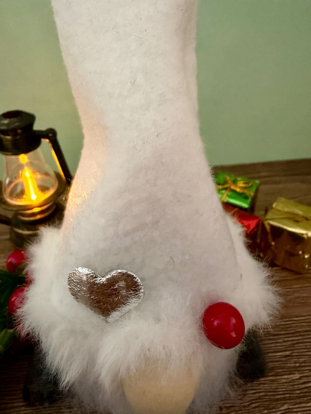 A close-up of a white hatted Christmas gnome decoration with fluffy trim, a silver heart embellishment, and a wooden bead nose, set on a wooden surface with holiday decor in the background.