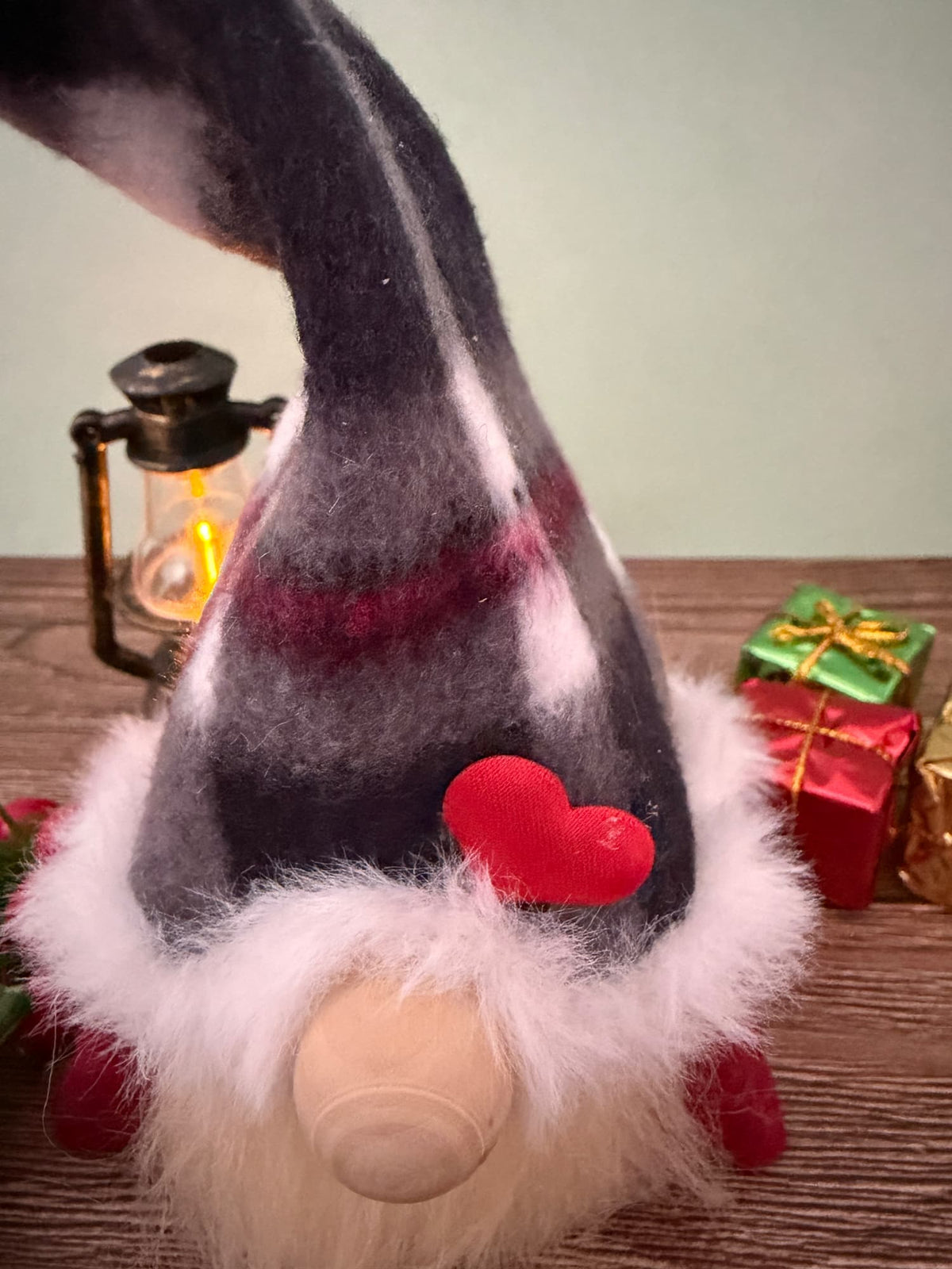 A close up of Christmas gnome with a dark gray and white striped hat, fluffy white beard, and red felt heart detail, displayed on a wooden surface with a lantern and wrapped gifts in the background.