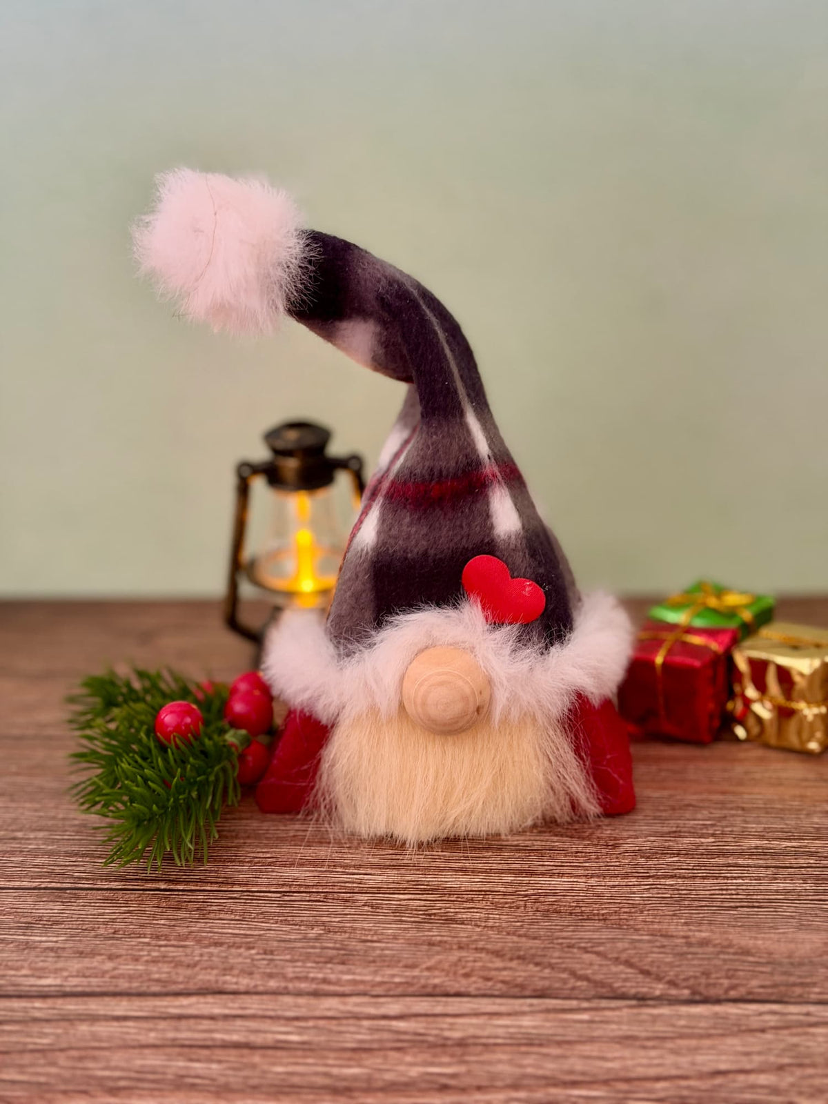 A handmade Christmas gnome figurine with a tall balck and red checked hat, fluffy blonde beard, and wooden nose sits on a wooden surface with a lit lantern, greenery, and wrapped gifts in the background. Made by Northern Nomes, part of the Yuletide folk collection.
