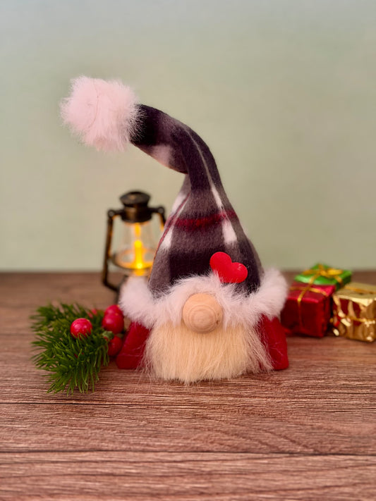 A handmade Christmas gnome figurine with a tall balck and red checked hat, fluffy blonde beard, and wooden nose sits on a wooden surface with a lit lantern, greenery, and wrapped gifts in the background. Made by Northern Nomes, part of the Yuletide folk collection.