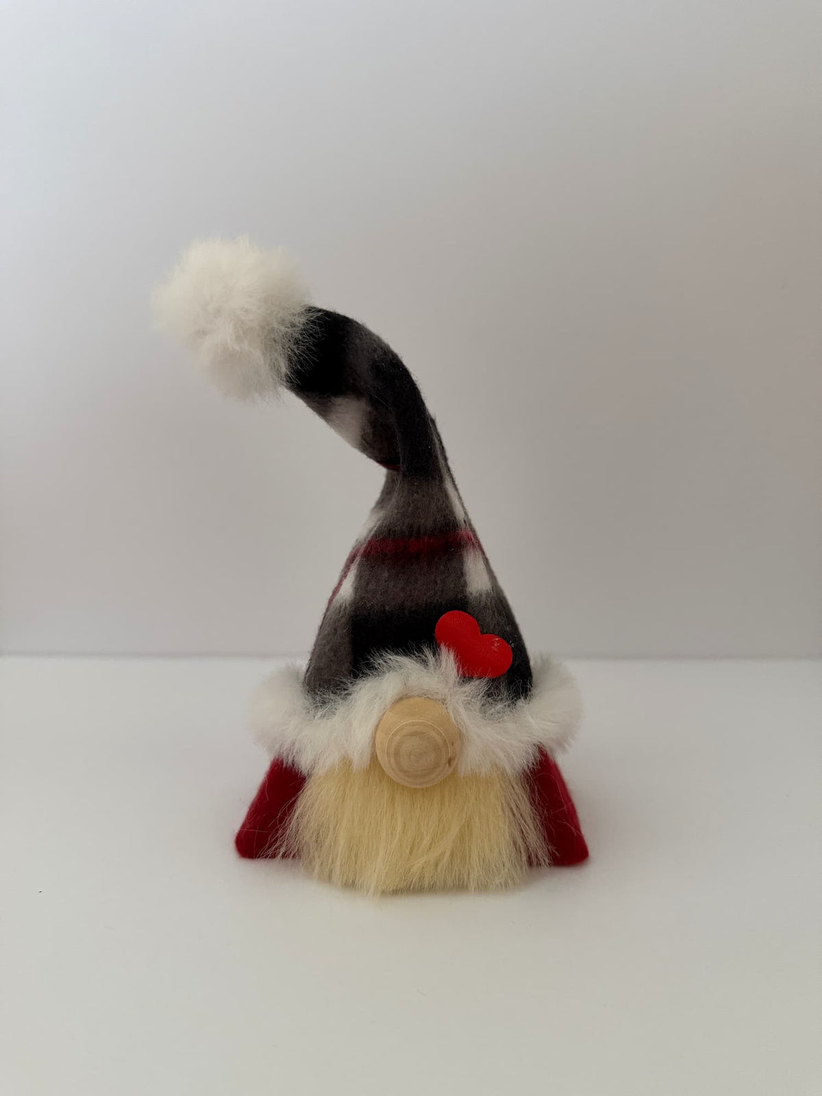A small Christmas gnome figurine with a tall red and black checked hat, fluffy blonde beard, and wooden nose sits on a wooden surface with a lit lantern, greenery, and wrapped gifts in the background. Handmade by Northern Nomes.