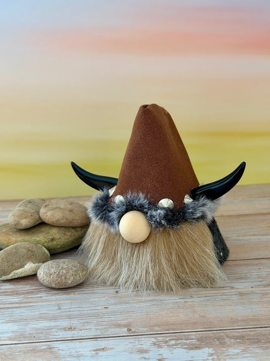 A small decorative handmade gonk figure  with a tall brown hat, black horn-like side pieces, a fluffy beige beard, and a gray faux-fur trim around the hat, displayed on a wooden surface with stones against a soft pastel background.