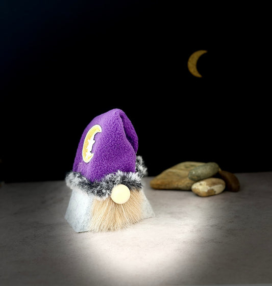 A small gnome figurine with a purple fleece hat, fluffy cream beard, grey faux-fur trim, and a round wooden nose, displayed on a tabletop with decorative stones and a crescent moon against a dark background. Handmade by Northern Nomes.