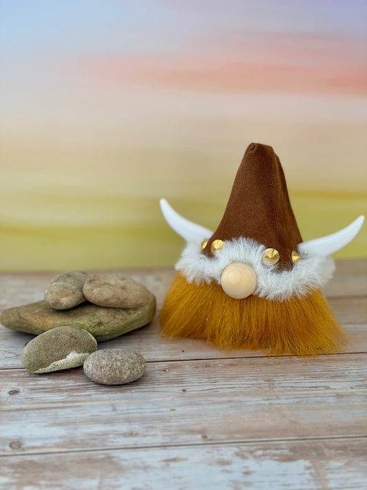 A small decorative gonk figurine styled like a Viking, with a brown pointed hat, white horn-like side pieces, a fluffy white and golden beard, and a round wooden nose, placed on a weathered wood surface beside stacked stones against a soft pastel gradient background. Handmade character gonk by Northern Nomes