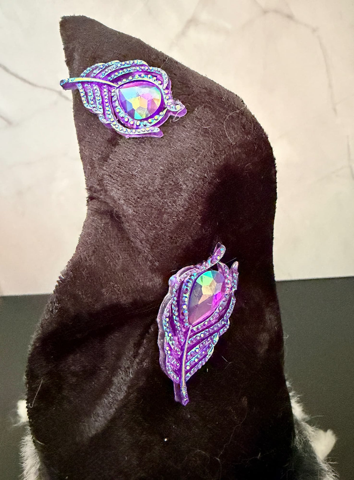 Two matching purple and iridescent leaf-shaped embellishments displayed on a dark black velvet witch gnome hat against a light marble-patterned background.
