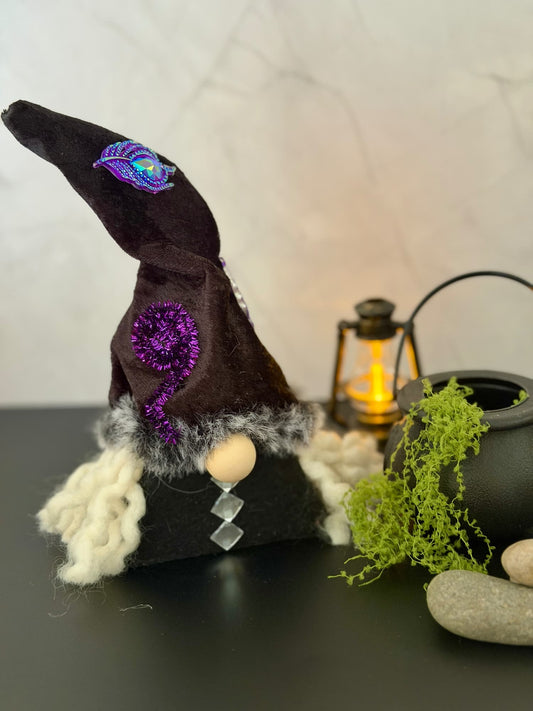 A decorative gnome witch figure with a tall dark black hat featuring purple embroidered details, white fuzzy hair and a black body, styled on a dark tabletop with a lantern, moss, and stones against a light marble wall.n Handmade by Northern Nomes.