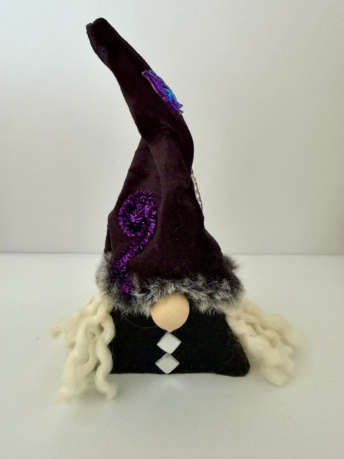 A small witch gnome with a tall dark black pointed hat, fluffy white yarn hair, a black body, and a round wooden bead nose, photographed against a plain light background.