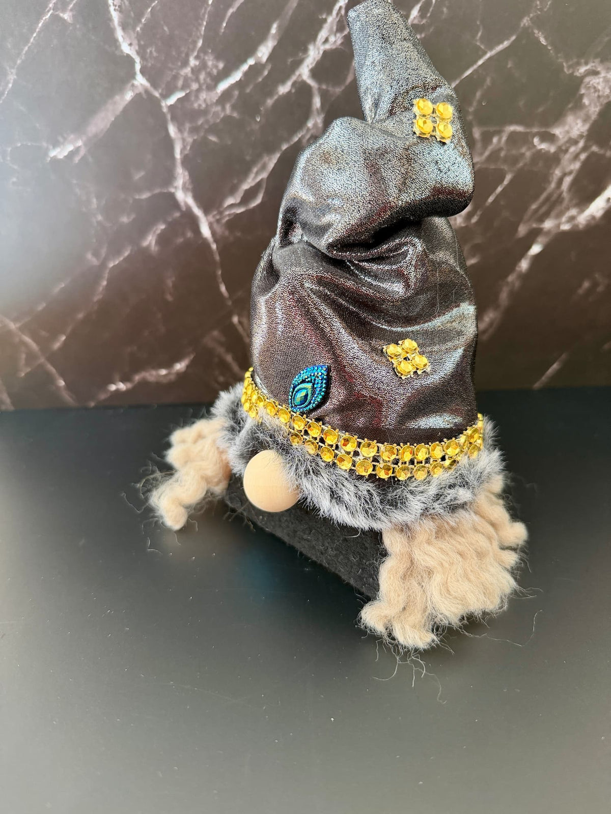 A small witch gnome figurine wearing a dark gray glittery pointed hat with gold embellishments, a blue eye detail, and a gold rhinestone trim, with beige yarn hair and a black base, set against a dark marble-patterned background.