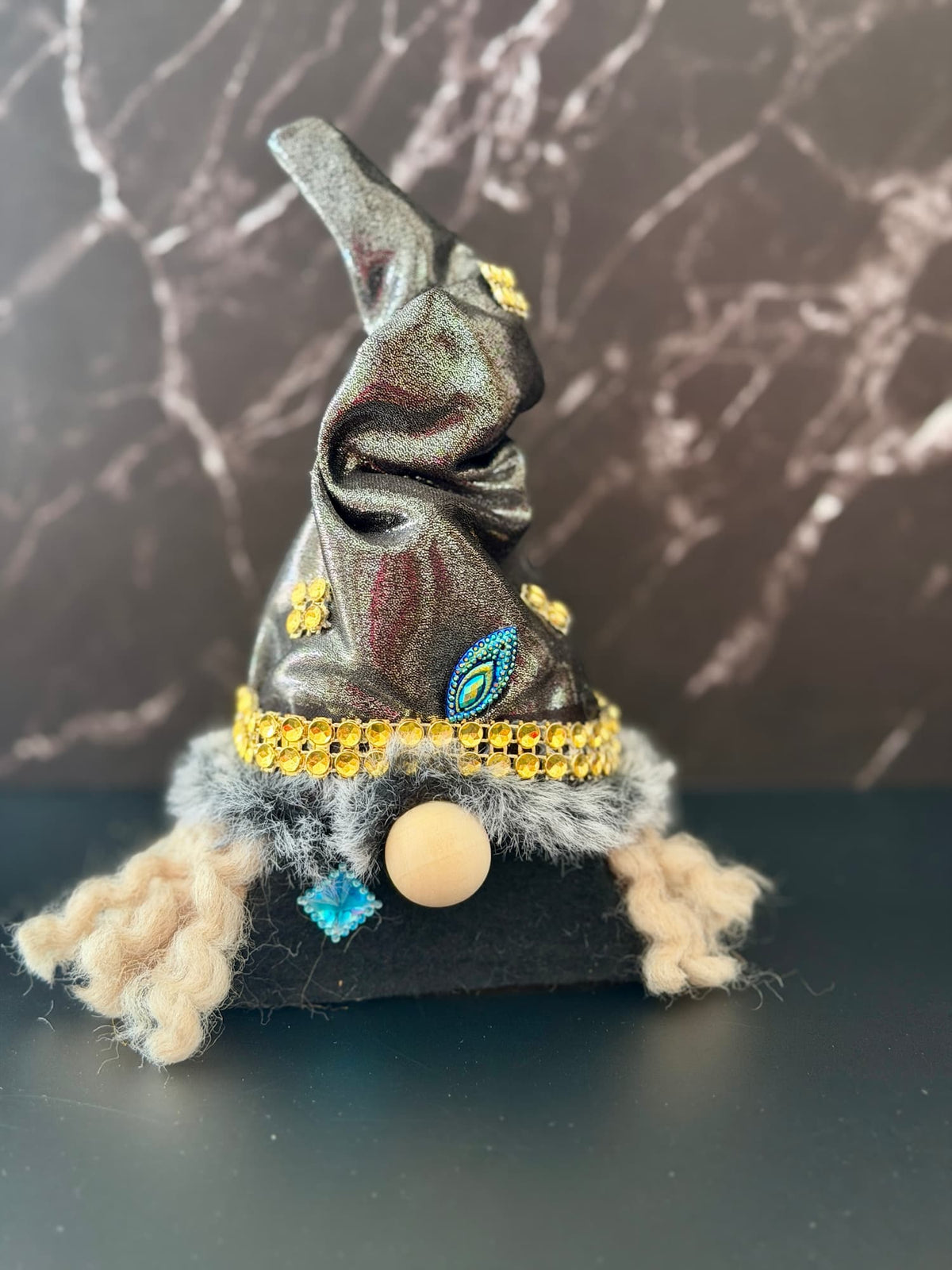 A small witch hat gnome decoration with a dark metallic gray hat, gold beaded band, fluffy gray trim, beige hair, and a round wooden nose, set against a dark marble-patterned background. Handmade by Northern Nomes.