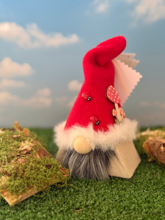 A small gnome figurine styled as a mushroom, wearing a red fleece hat with white faux-fur trim, standing on artificial green grass against a blue sky backdrop with clouds. The gnome has grey faux fur, a beige wooden nose, and decorative red and pink mushroom embellishments on the hat. Handmade by Northern Nomes and part of the Mosswood collection of Wee Folk.