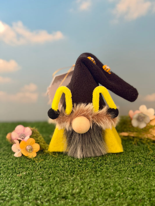 A handmade gnome styled like a bee, featuring a black hat with yellow antennae, a fuzzy yellow body, and a round beige wooden nose, set on artificial green grass with small flowers and a blue sky backdrop. Made by Northern Nomes
