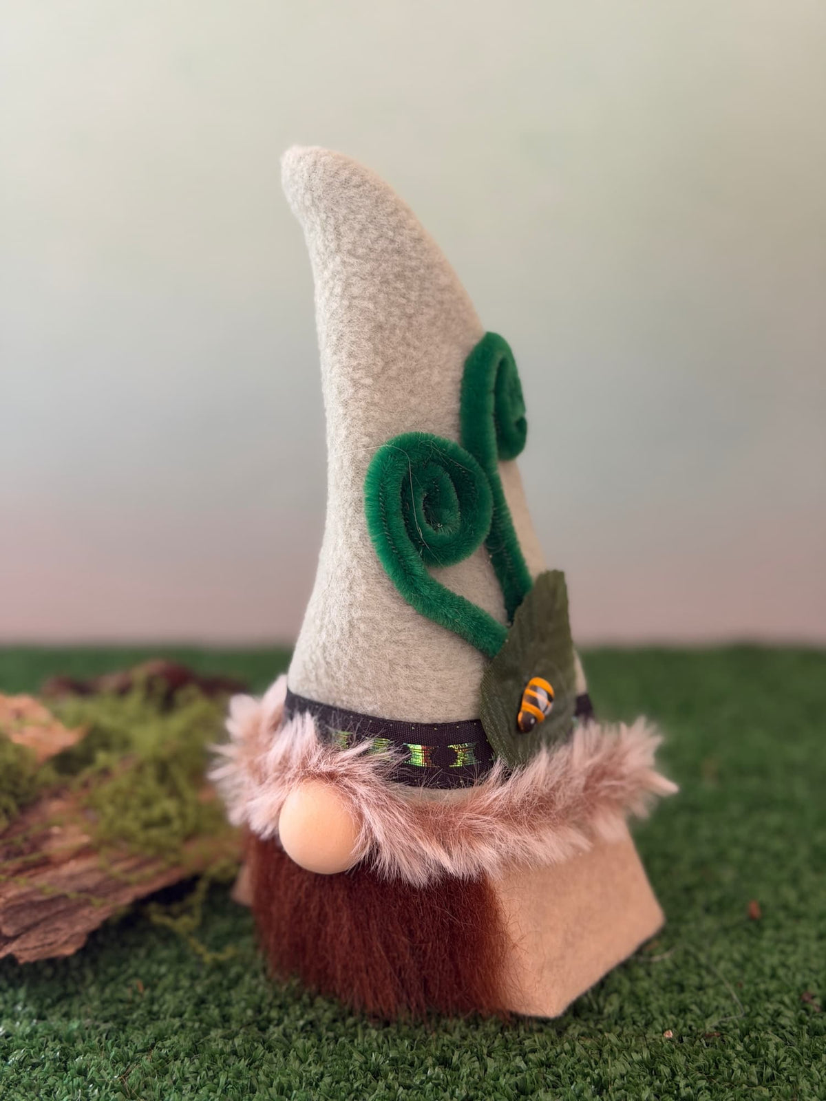 A handmade gnome with a tall greenish hat decorated with green felt swirls and a small leaf, standing on artificial green grass with a blurred background.