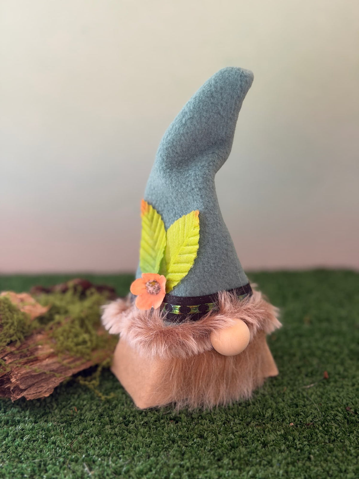 A handmade gnome figurine with a tall light greenish hat decorated with green leaves and an orange flower, a tan beard, and a small wooden nose, set on artificial grass with a mossy log prop.