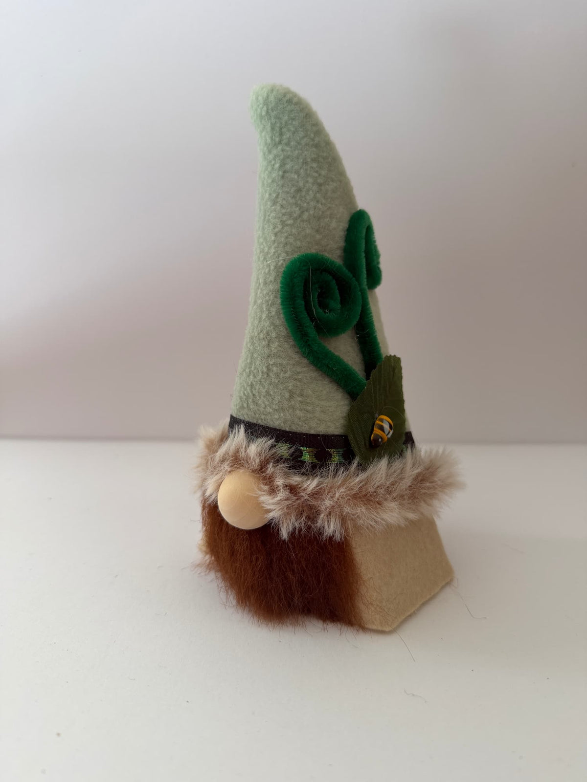 A small handmade nome decoration with a pointed light green hat, dark green leaf detail, brown faux-fur trim, a beige wooden nose, and a tan body base, photographed against a plain light background. Made by Northern Nomes.