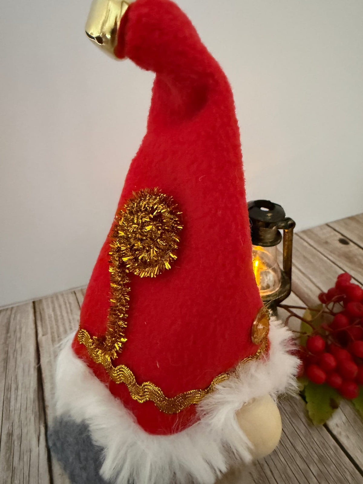 A festive red elf gnome hat decoration with white faux fur trim, gold tinsel accents, and a gold jingle bell tip, displayed on a rustic wooden surface next to a lantern and red berries.
