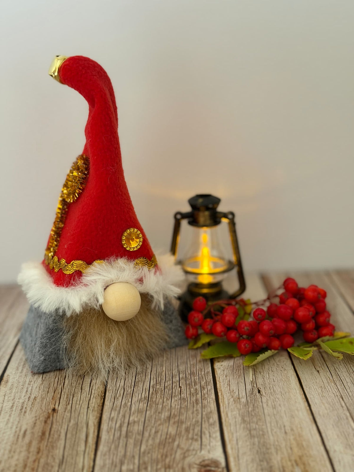 A handmade festive Christmas elf gnome figurine by Northern Nomes with a tall red hat trimmed in white faux fur and gold glitter embellishment,standing on a rustic wooden surface next to a lit vintage lantern and red. berries. 