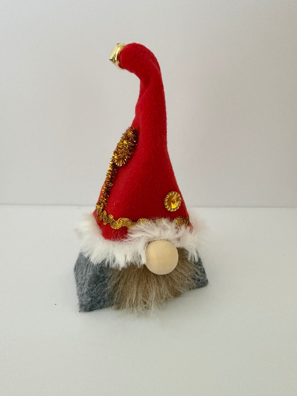 A small Christmas elf gnome decoration with a tall red felt hat trimmed in gold sequins, white faux fur, and a gray felt body, standing on a white surface against a white background. Handmade by Northern Nomes.
