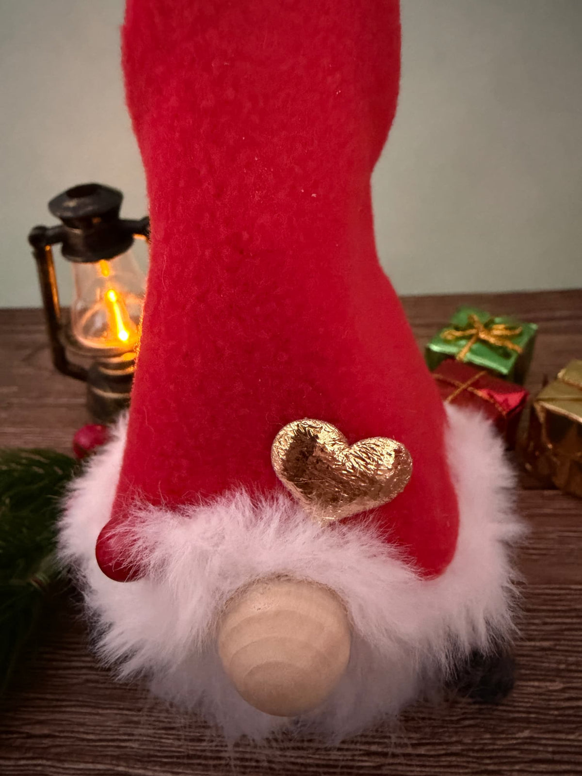 A festive Christmas gnome decoration with a tall red felt hat, fluffy white faux fur trim, a small gold glitter heart, and a round wooden nose, displayed on a wooden surface with holiday props in the background.