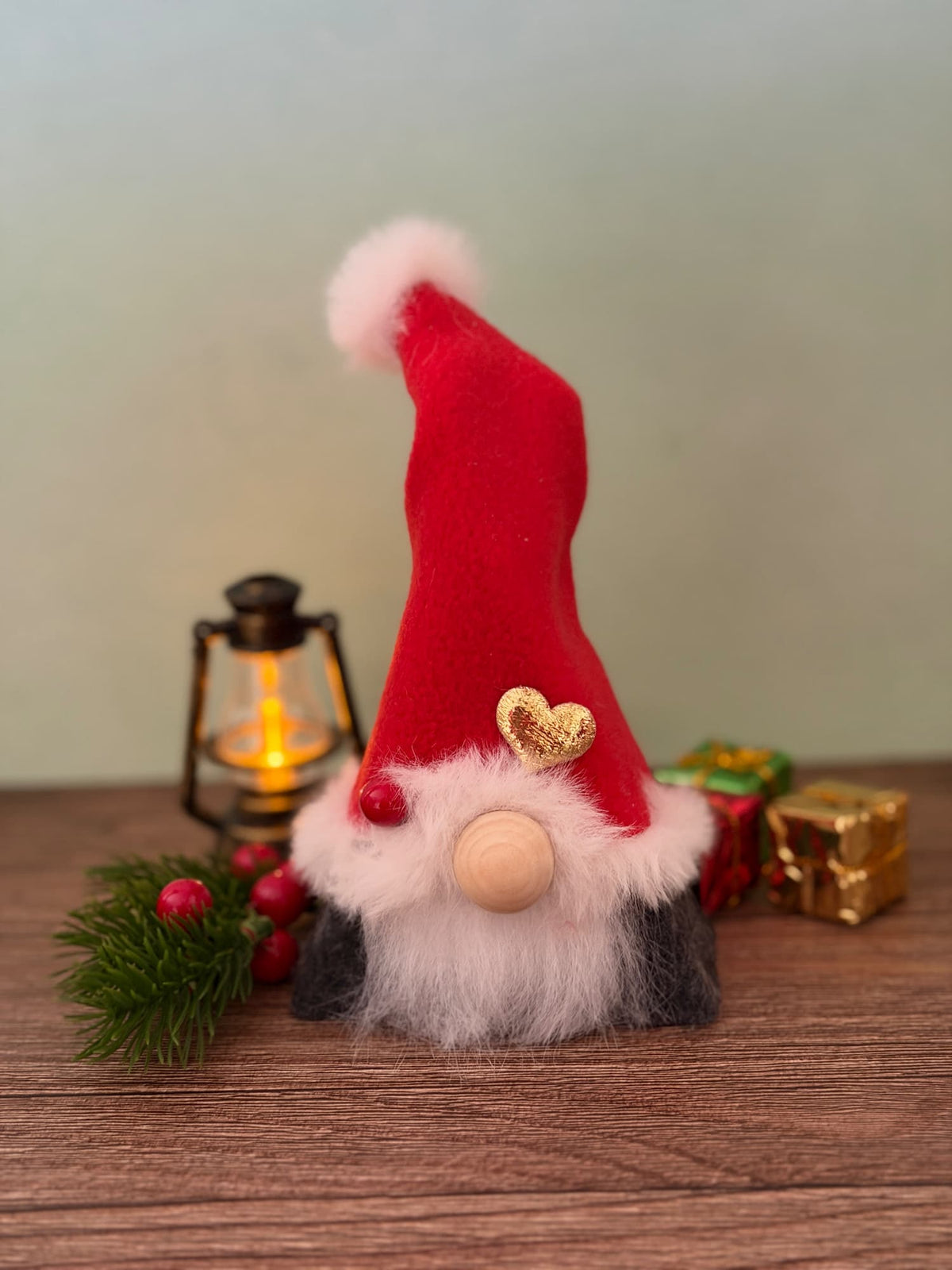 A small Christmas gnome figurine with a tall red hat, fluffy white beard, and wooden nose sits on a wooden surface with a lit lantern, greenery, and wrapped gifts in the background. Handmade by Northern Nomes, part of the Yuletide Folk collection.