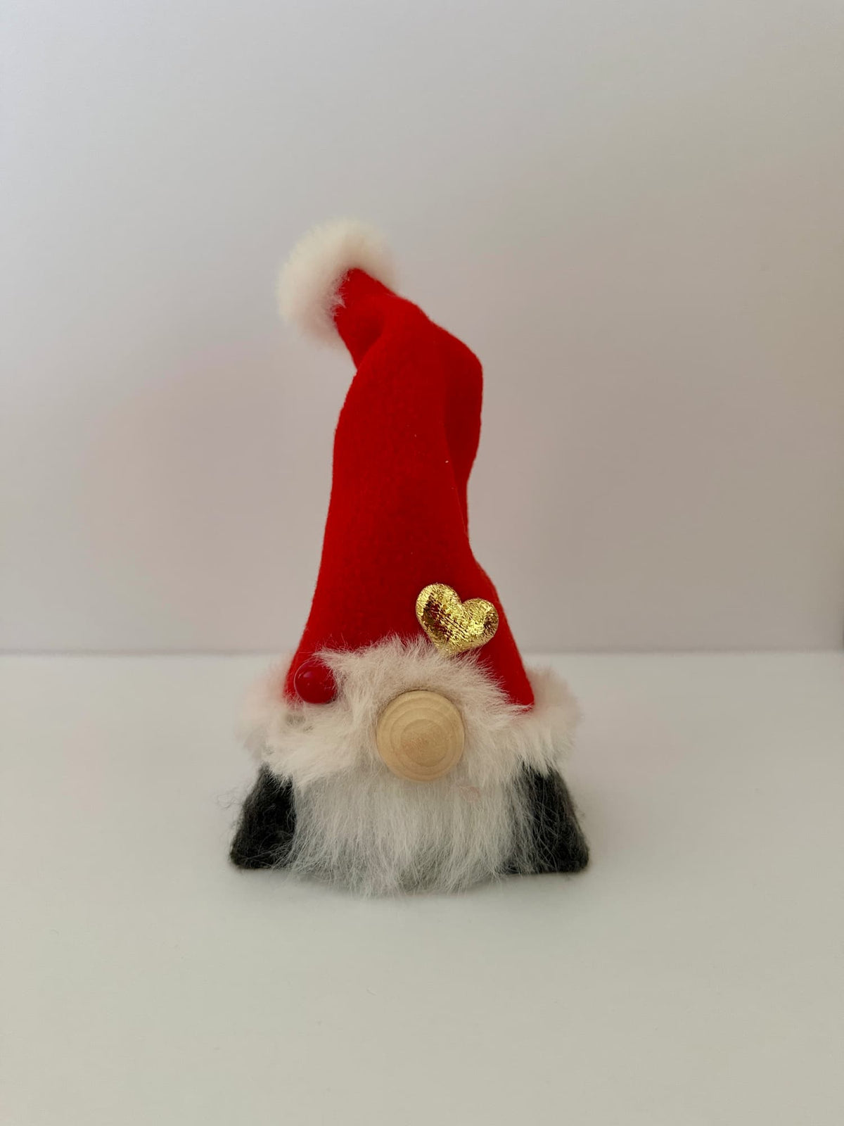 A handmade small gnome decoration with a tall red hat, white beard, dark grey felt covered body, a wooden button nose, and a gold heart ornament, standing on a white surface against a light gray background. Made by Northern Nomes.