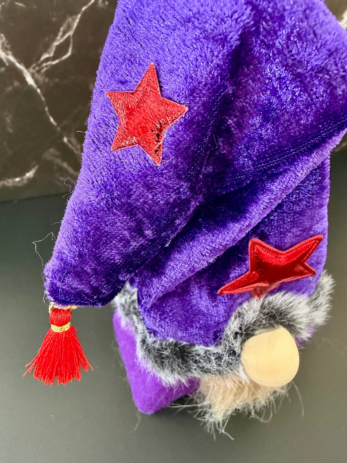 A close-up of a purple plush wizard hat with red embroidered stars, a red tassel, and a gray faux-fur trim, shown against a dark background.