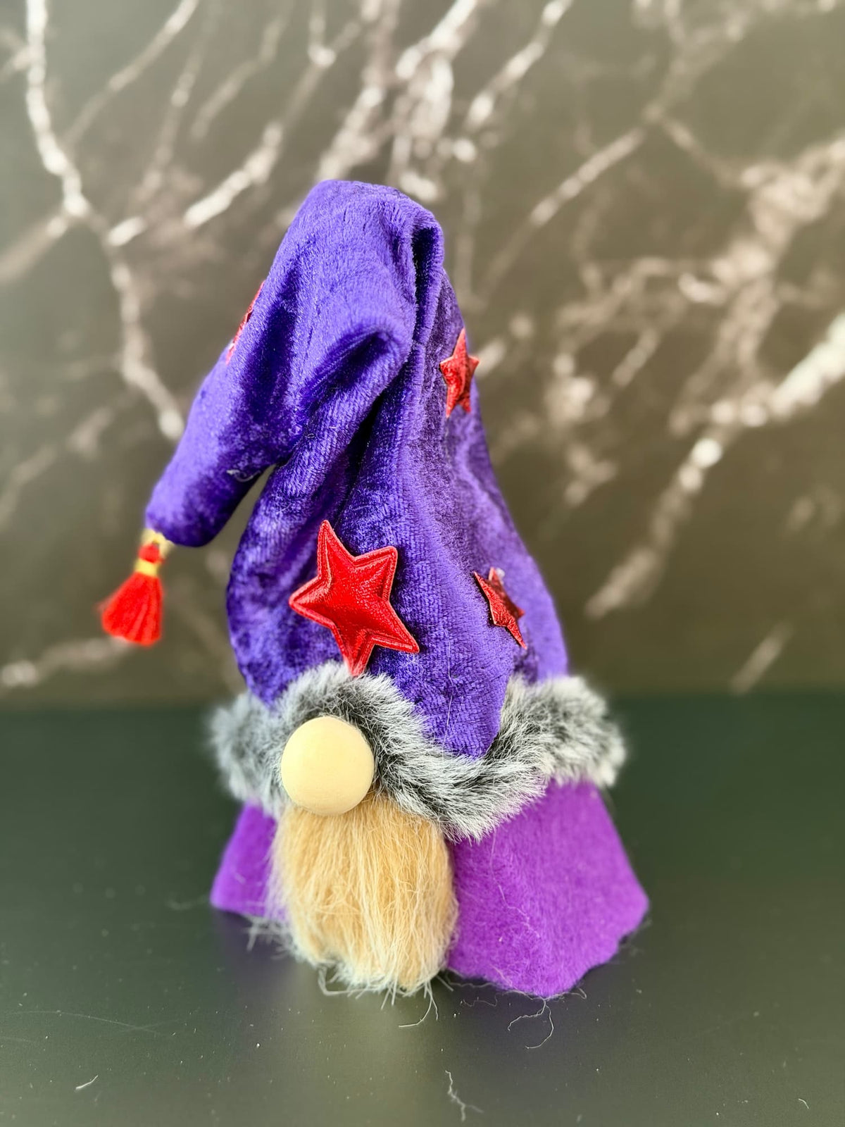 A small felt wizard gnome figurine wearing a purple pointed hat with red star appliqués and a fluffy cream beard, standing on a dark surface against a gray marble-patterned background. Handmade by Northern Nomes.