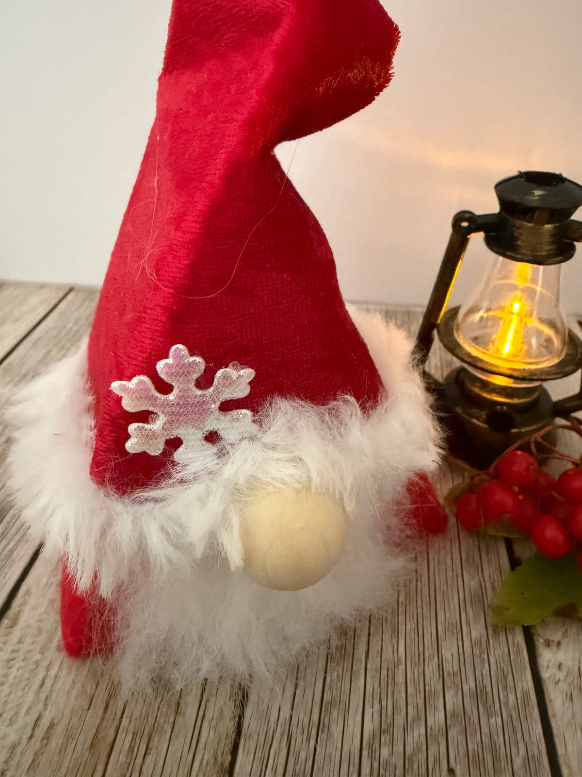 A festive Christmas gnome decoration with a red felt hat, fluffy white faux fur trim, a white snowflake ornament, and a round wooden nose, styled on a rustic wooden surface next to a lit vintage lantern.