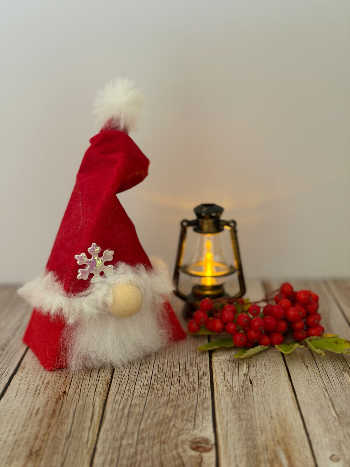 A festive red felt gnome decoration wearing a Santa-style hat with white faux fur trim, a wooden beanie, and a silver snowflake ornament, displayed on a rustic wooden surface beside a lit lantern and red berries. Handmade by Northern Nomes.