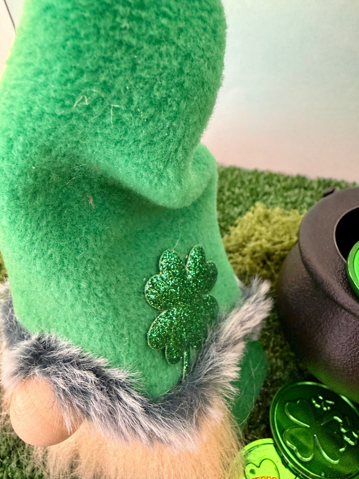 A close-up of a handmade green felt St. Patrick's Day gnome decoration with a glittery green shamrock patch, faux fur trim, and a beige wooden nose, shown next to a black pot and green shamrock coins.