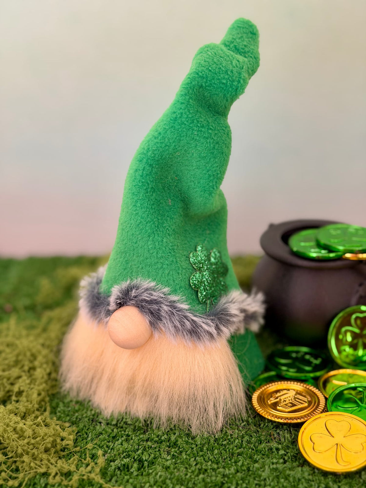 A St. Patrick's Day lucky Leprechaun gnome with a tall green hat, fluffy cream beard, and green faux fur trim, styled on green moss next to a black pot of coins and gold shamrock coins. Handmade by Northern Nomes.
