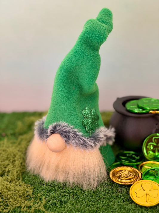 A St. Patrick's Day lucky Leprechaun gnome with a tall green hat, fluffy cream beard, and green faux fur trim, styled on green moss next to a black pot of coins and gold shamrock coins. Handmade by Northern Nomes.