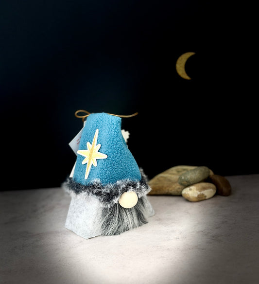 A small gnome decoration with a blue pointed hat, yellow star detail, grey faux-fur trim, and a round wooden nose, standing on a light surface against a dark background with a crescent moon and stones. Handmade by Northern Nomes, part of the Moonshadow Folk collection.